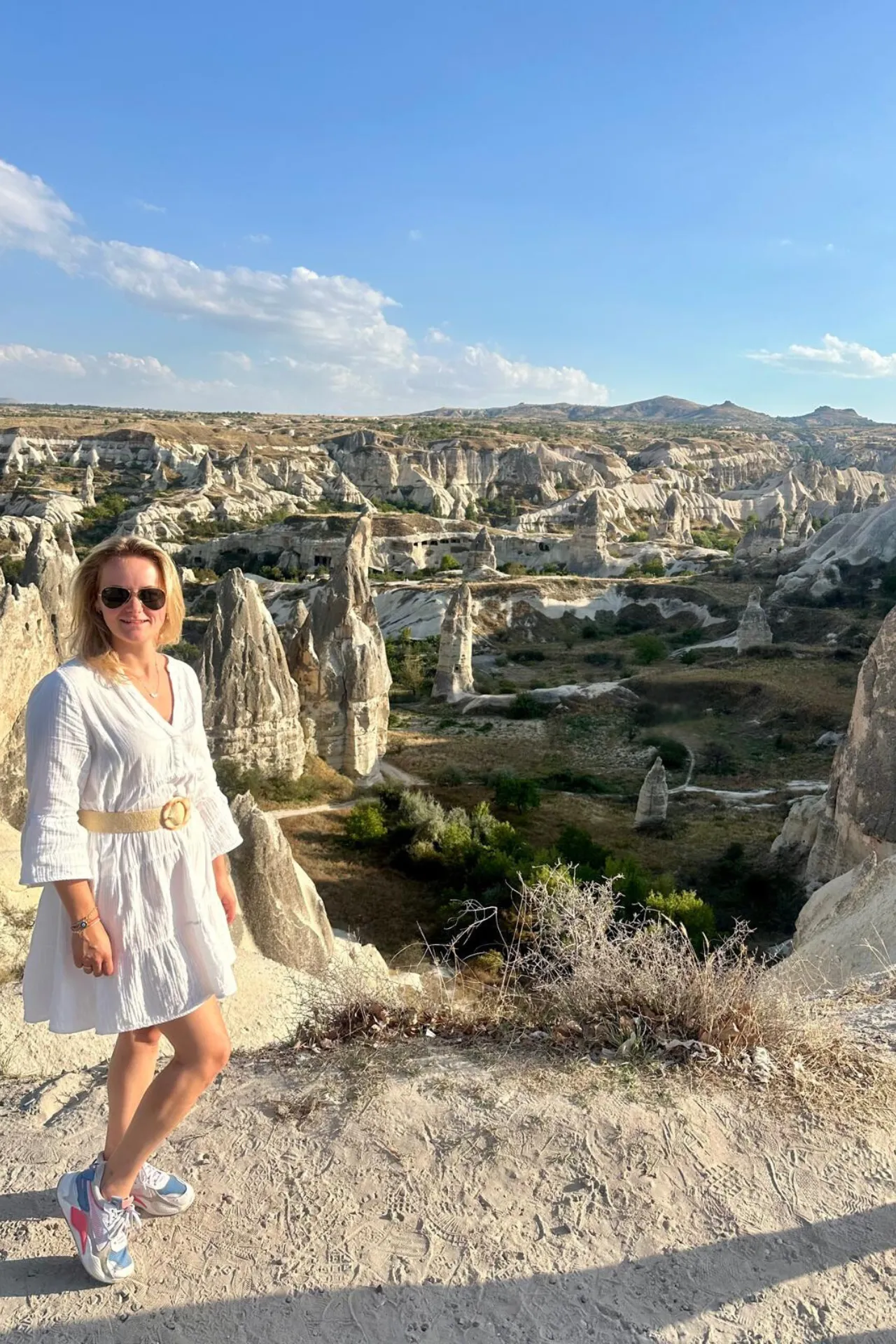 Persoon staat op een rotsplateau met uitzicht over het spectaculaire landschap van Cappadocië, Turkije. Op de achtergrond zijn grillige rotsformaties, valleien en uitgehouwen grotten zichtbaar, typerend voor deze historische regio die bekendstaat om zijn unieke geologie en culturele erfgoed. De heldere blauwe lucht en het zonlicht benadrukken de natuurlijke kleuren van het gesteente. Deze locatie is een populaire bestemming voor toeristen en fotografen die op zoek zijn naar bijzondere bezienswaardigheden en panoramische uitzichten.