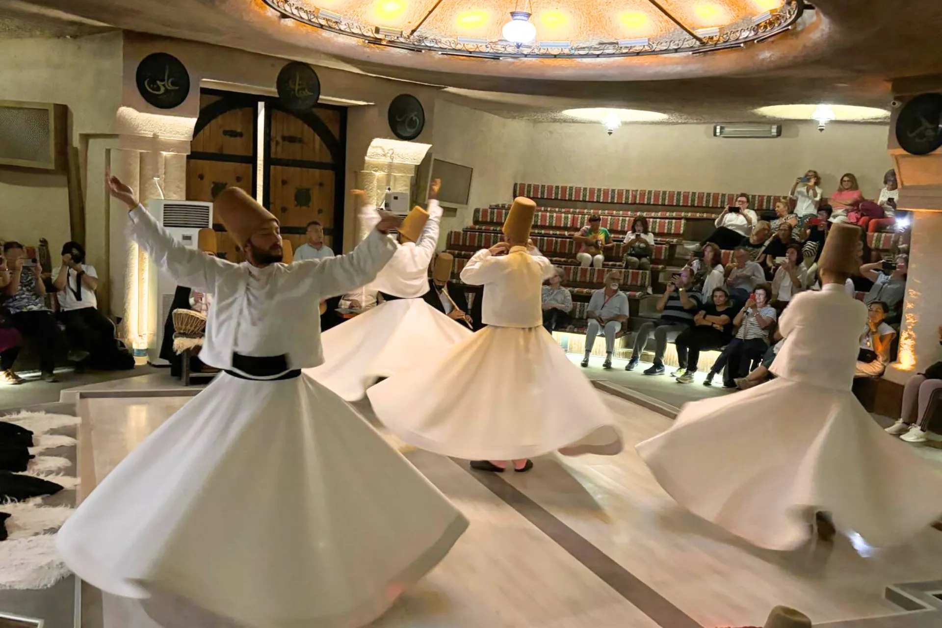 Traditionele Sufi-ceremonie met draaiende derwisjen in Cappadocië, Turkije. Vier dansers in witte gewaden en hoge hoeden voeren de spirituele dans uit op een ronde houten vloer, terwijl het publiek op verhoogde zitplaatsen toekijkt. Het plafond is versierd met warme verlichting en islamitische kalligrafie, wat een authentieke en serene sfeer creëert. Deze eeuwenoude rituele dans, bekend als de Semazen, is een belangrijk cultureel erfgoed en een populaire attractie voor toeristen die Turkse tradities willen ervaren.