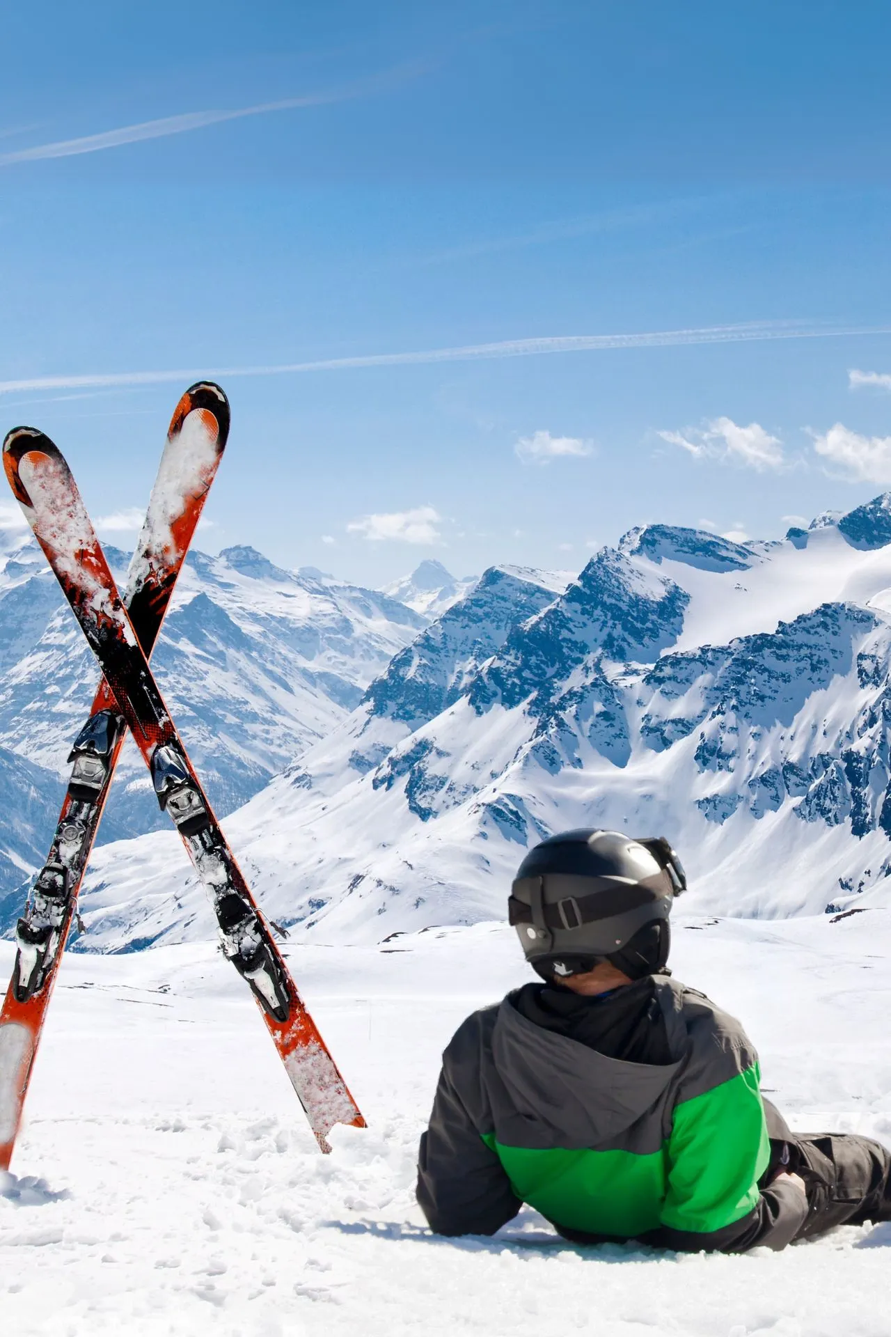 Skiër met helm en groene jas zit in de sneeuw en kijkt uit over een adembenemend berglandschap met besneeuwde toppen