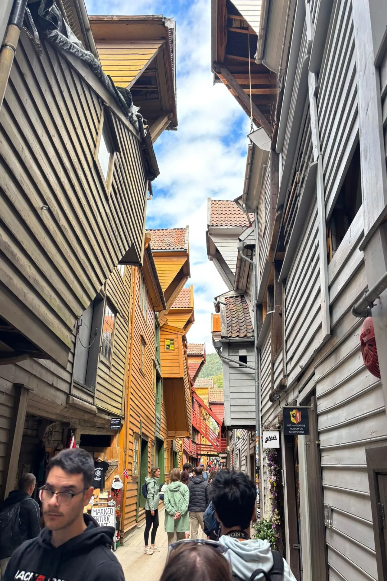 Smalle steeg met hoge, houten gebouwen in verschillende kleuren zoals grijs, bruin en oranje, met overhangende daken en houten balkons. Mensen lopen door de straat, die een historische en pittoreske uitstraling heeft. De lucht is deels bewolkt met blauwe stukken zichtbaar.