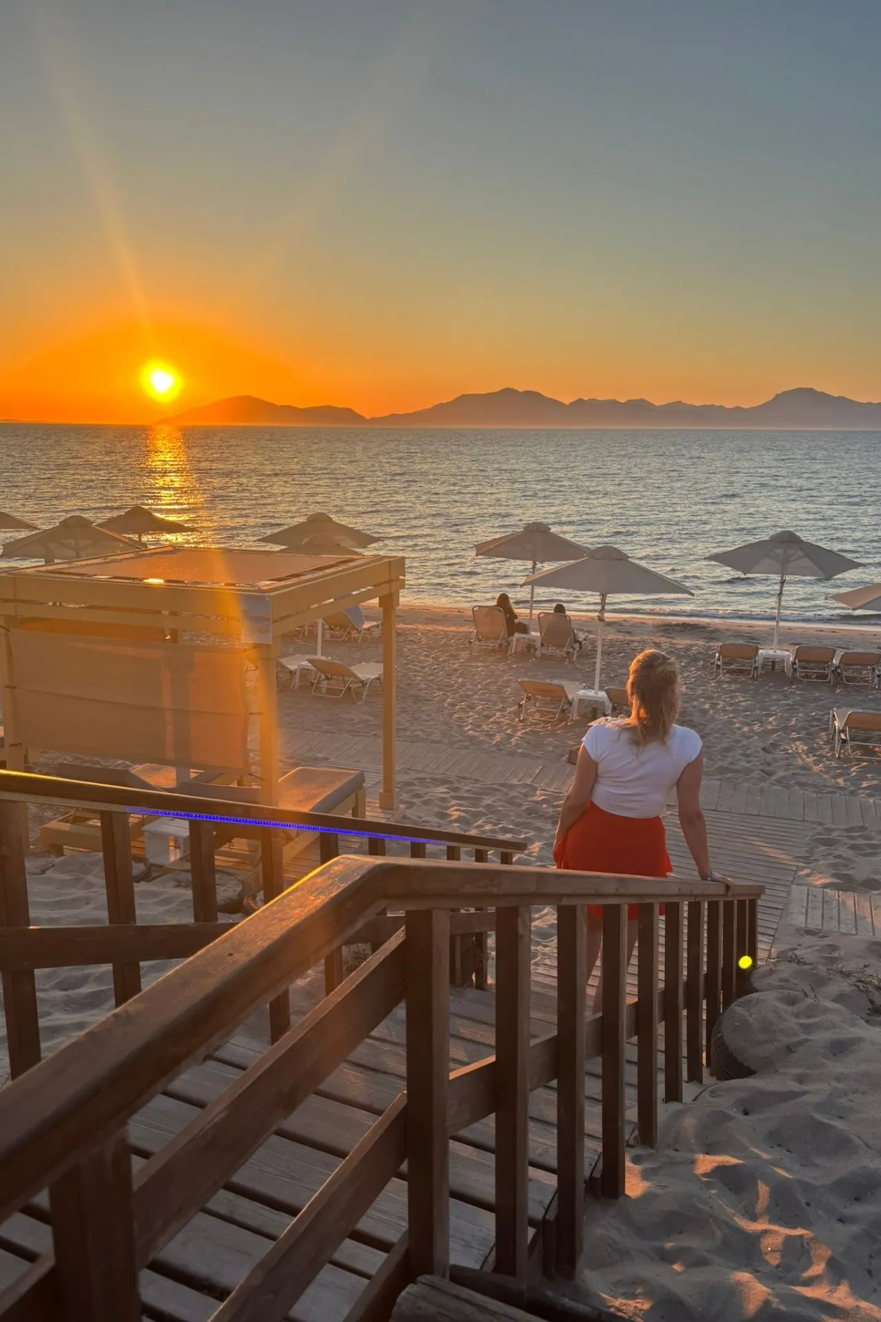 Vrouw geniet van een betoverende zonsondergang boven de Egeïsche Zee vanaf een houten trap bij het strand, met ligbedden en parasols op de voorgrond.