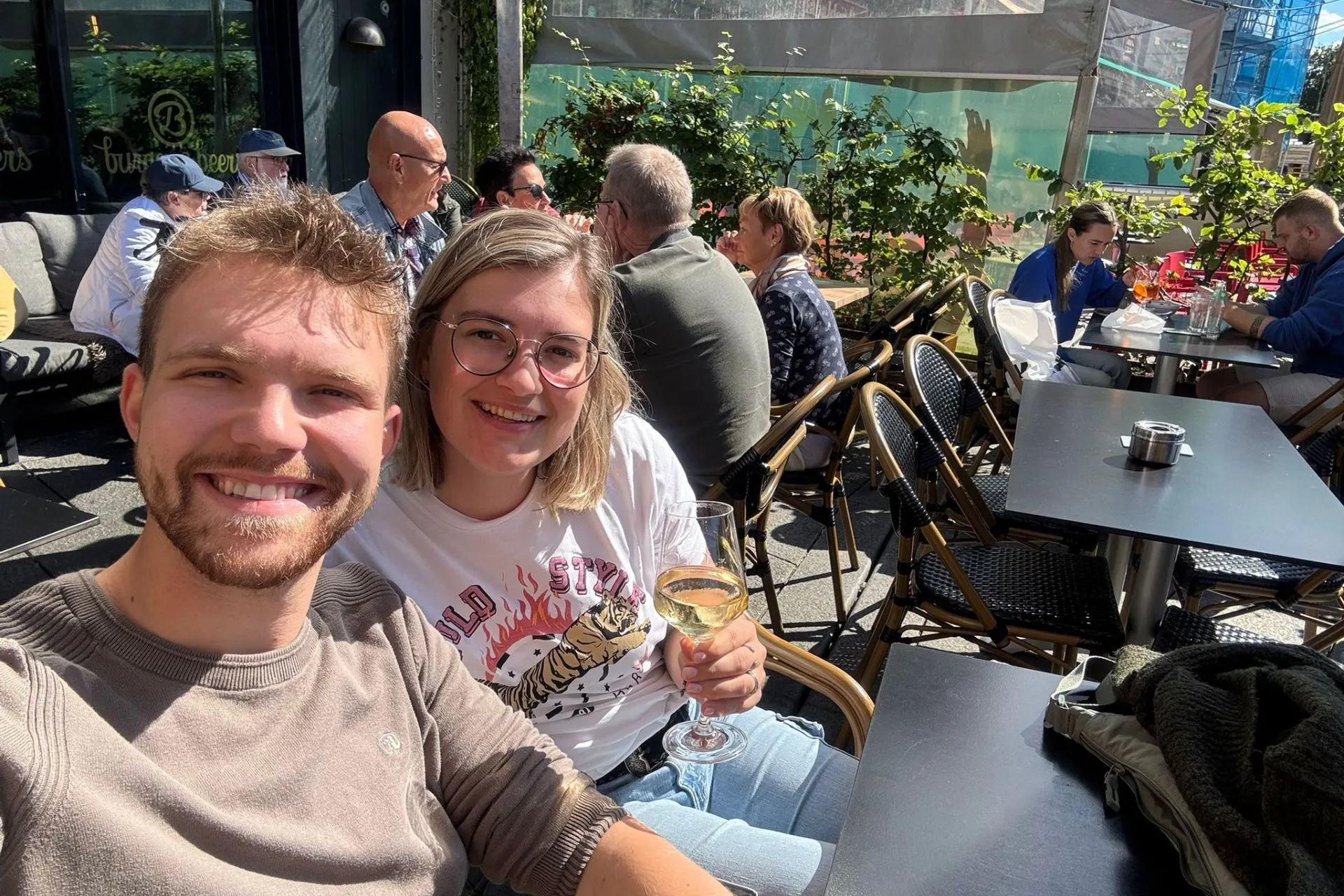 Twee personen zitten op een buitenterras van een café of restaurant, omringd door andere gasten aan tafels. Op de tafel staat een glas witte wijn en een asbak, met stoelen en tafels in een zonnige setting. Achtergrond toont groene planten en een gedeeltelijk afgedekte bouwplaats.