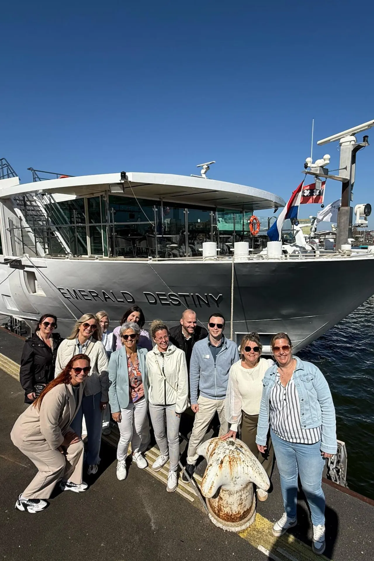 Groepsfoto van lachende deelnemers aan boord van de Emerald Destiny, met de riviercruiseboot op de achtergrond op een zonnige dag.