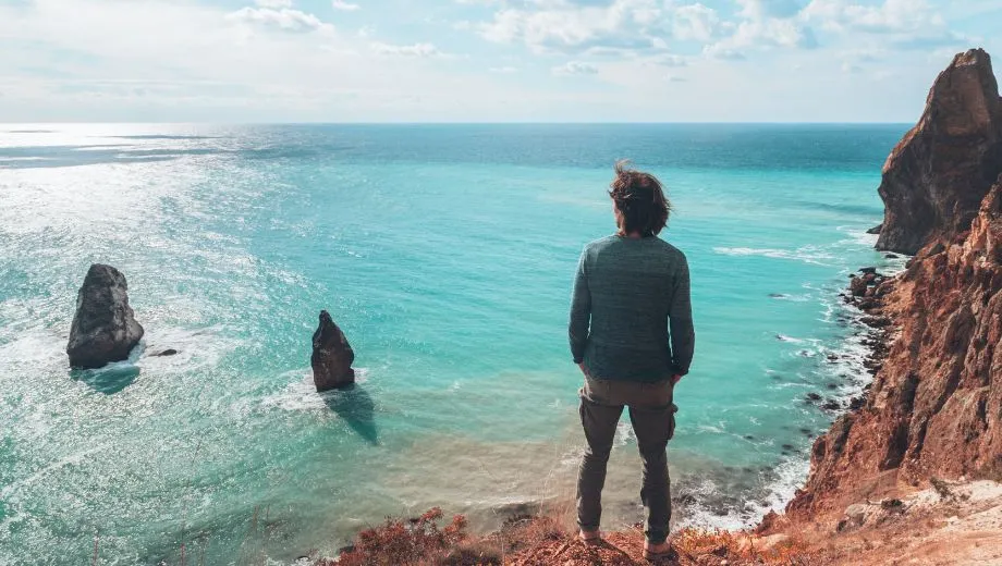 Een man staat met zijn handen in zijn zakken op een rotsachtig strand en kijkt uit over een blauwe zee. De wind waait door zijn haar onder een licht bewolkte, blauwe lucht. Het weer oogt mild, niet heet, en de man draagt een trui voor extra warmte.
