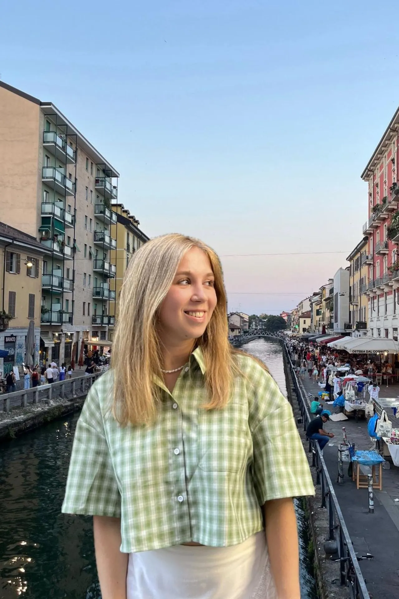 Marketeer Martje poseert lachend aan het kanaal van Navigli in Milaan, met op de achtergrond kleurrijke gebouwen, terrasjes en een levendige avondmarkt langs het water.
