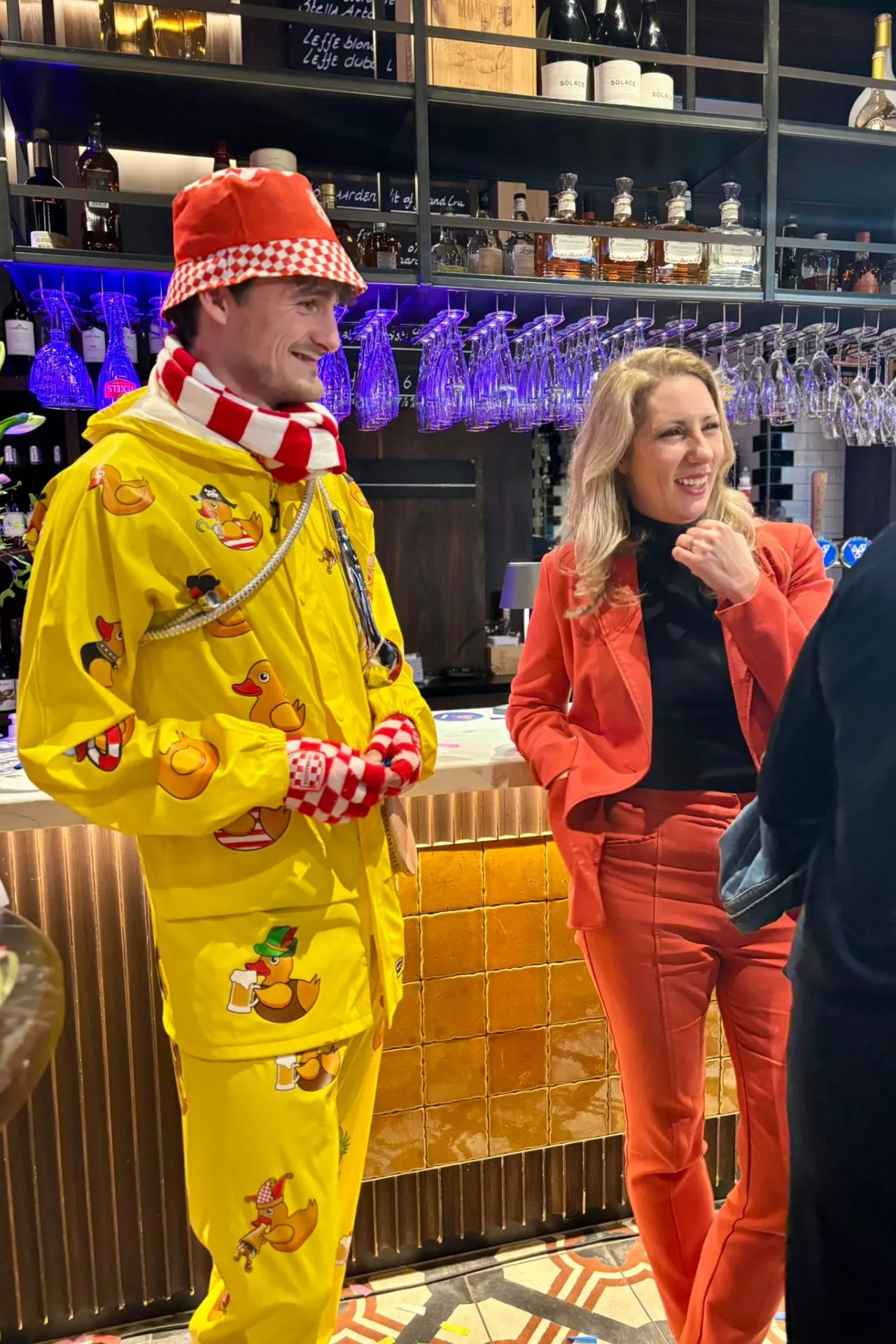 Levendige barsetting tijdens carnaval met een opvallende gele outfit vol eendjes en een rood-wit geblokte sjaal naast een stijlvolle outfit in rood. Op de achtergrond een goed gevulde bar met glazen en flessen, sfeervolle verlichting en warme kleuren. Deze foto straalt feestelijke Brabantse gezelligheid uit, ideaal voor carnavalsborrels, netwerken en sociale evenementen in een moderne horecalocatie.