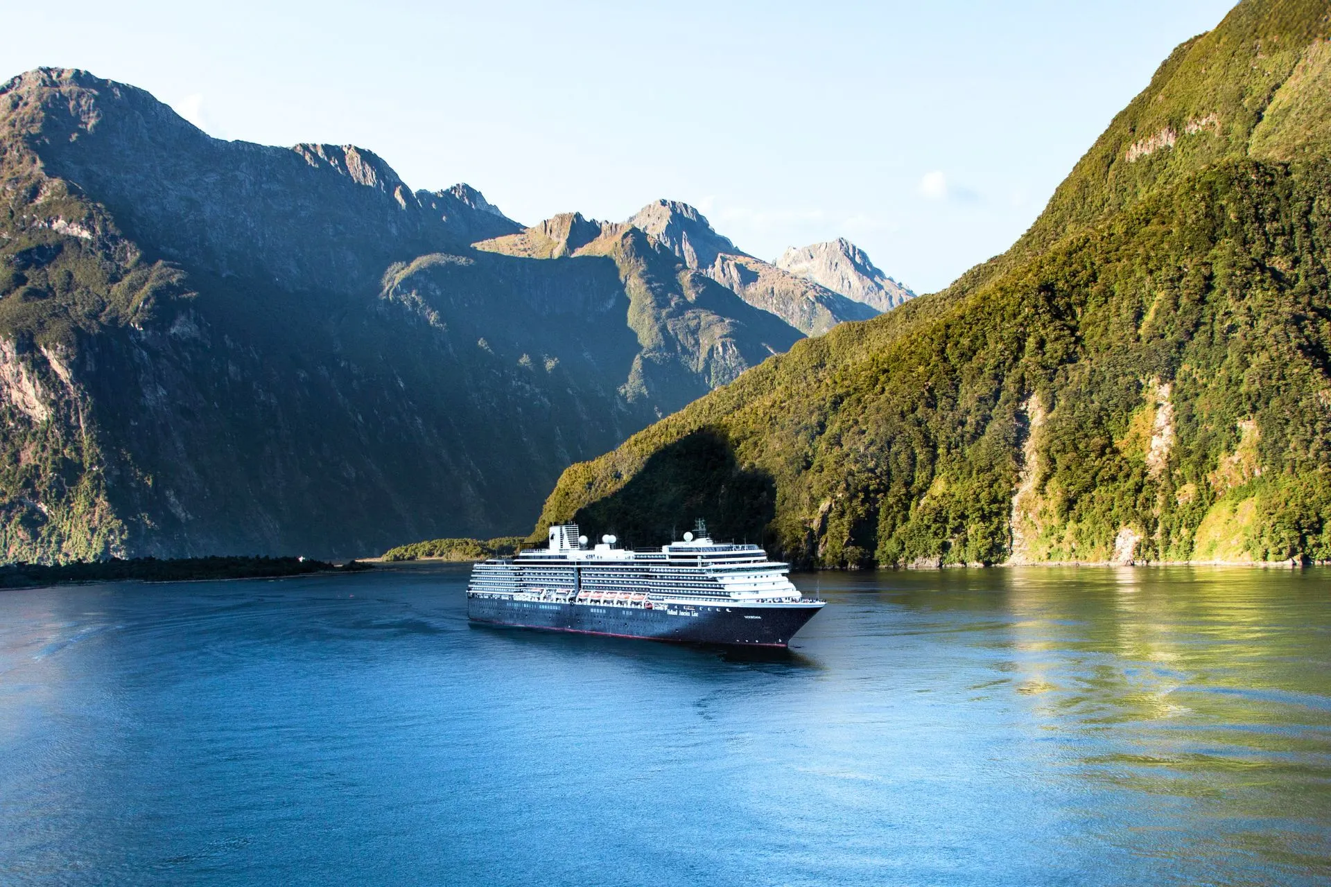 Cruiseschip van Holland America Line vaart door een indrukwekkende fjord omringd door steile bergen en dichtbegroeide hellingen. Het rustige water weerspiegelt het groene landschap, terwijl het schip door een van de mooiste natuurgebieden van Nieuw-Zeeland navigeert. Ideaal beeld van een luxe cruisevakantie met Holland America Line door spectaculaire fjorden en ongerepte natuur.