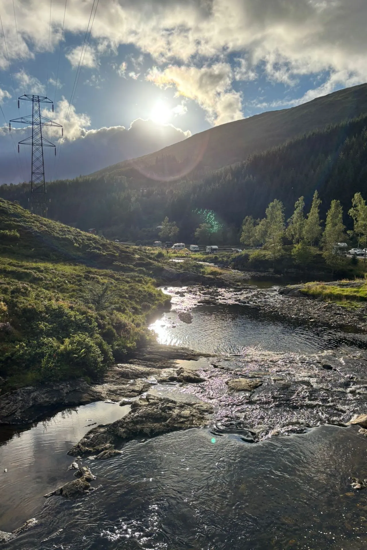 Zonnig berglandschap met een kabbelende beek op de voorgrond, omringd door groene heuvels en bomen. Op de achtergrond staan enkele campers en een hoogspanningsmast, terwijl de zon laag achter de berg schijnt door deels bewolkte lucht.