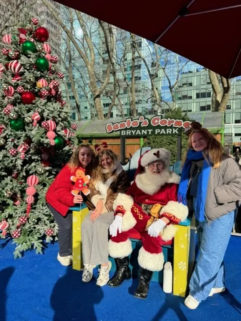Drie vrouwen poseren samen met de Kerstman in het feestelijk versierde Bryant Park in New York, omringd door kerstlichtjes en een winterse sfeer.