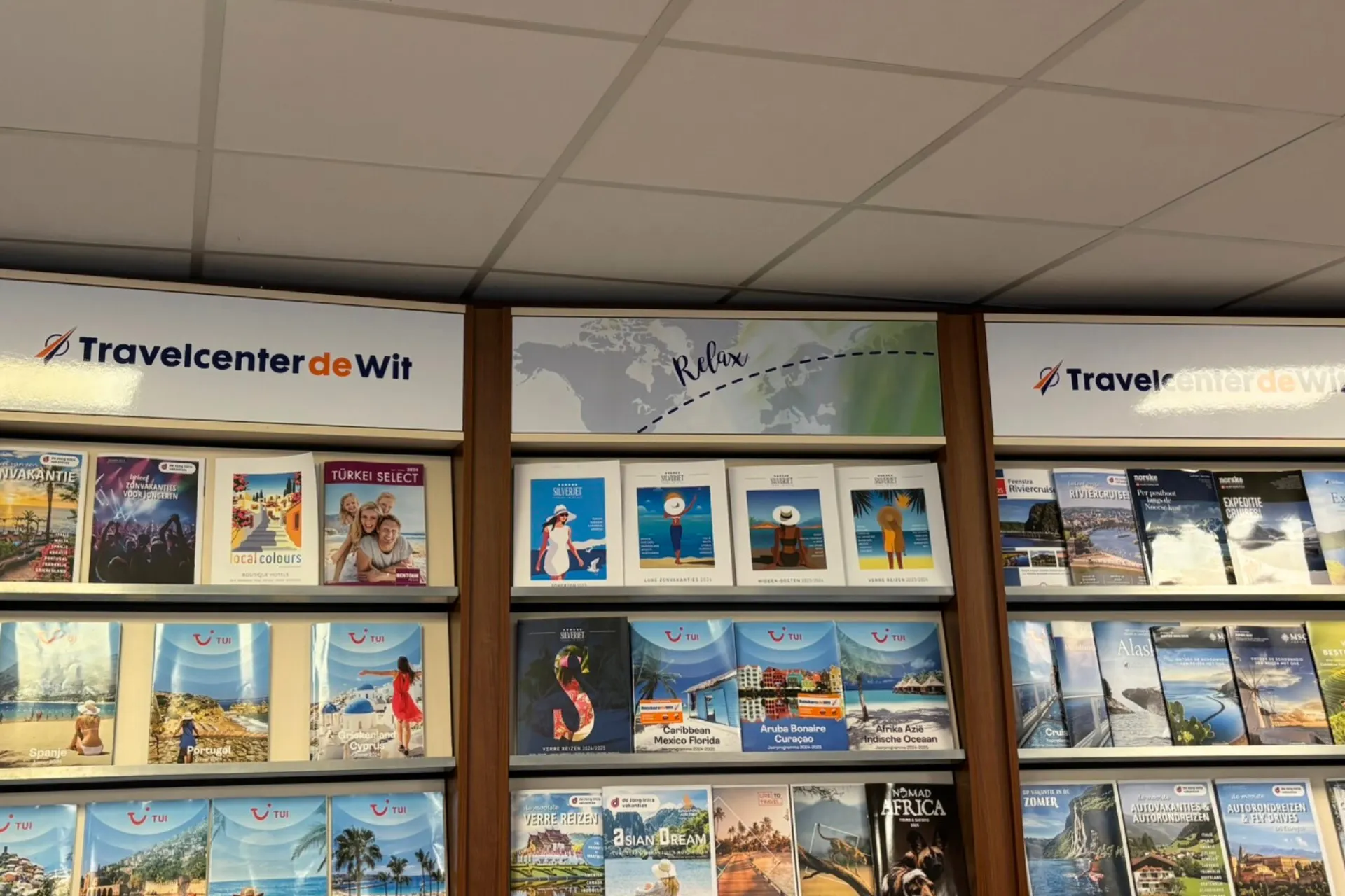 Wand met diverse reisboekjes van onder andere TUI, FOX en andere reisaanbieders, netjes uitgestald in rekken.