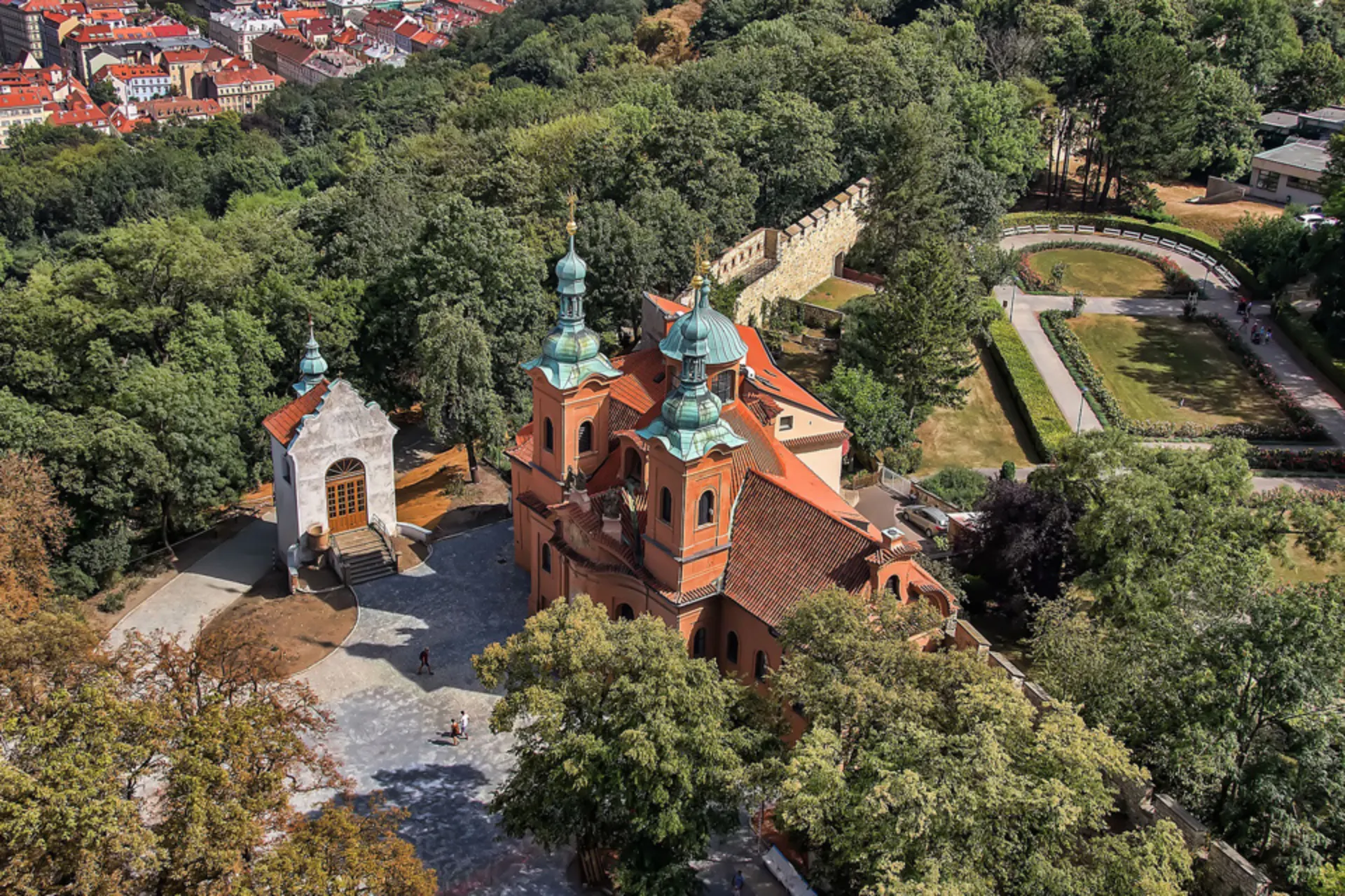 Luchtfoto van de historische Sint-Laurentiuskerk op de Petřín-heuvel in Praag, omringd door groene bomen en een park. De kerk heeft rode bakstenen muren en opvallende groene koperen koepels in barokstijl. Op de achtergrond zijn delen van de oude stadsmuur en rode daken van Praag zichtbaar. Populaire toeristische bezienswaardigheid in Tsjechië, ideaal voor cultuur, architectuur en sightseeing.