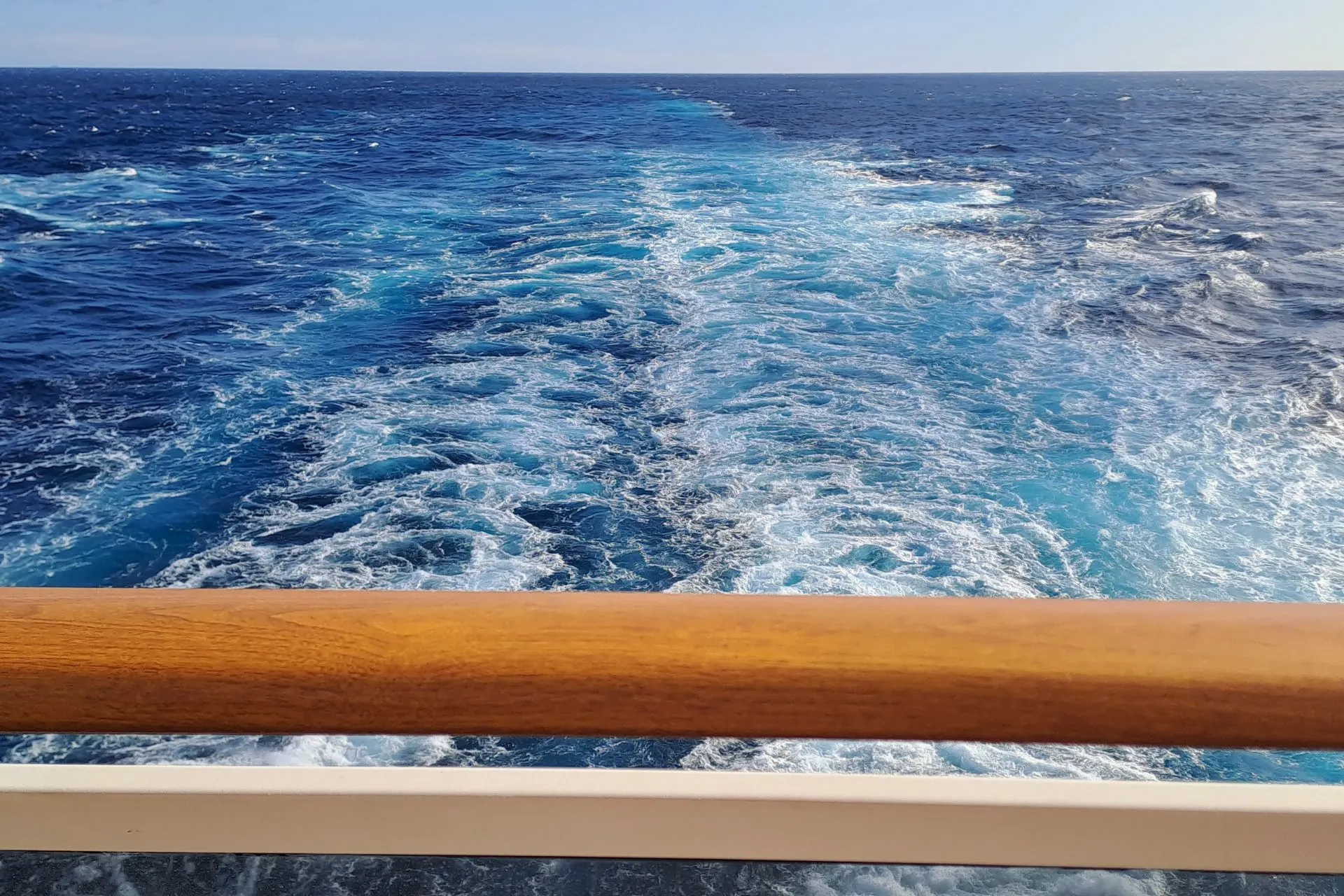 Achterkant van het cruiseschip, over een houten reling uitkijkend over het blauwe, golvende water.