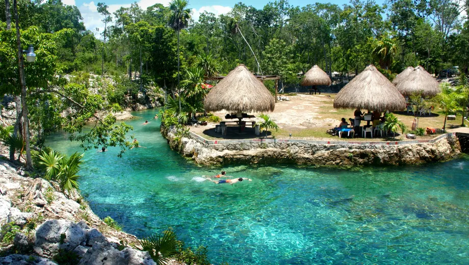 Natuurlijke cenote in een tropische omgeving met helder turquoise water, omringd door rotsen, groene vegetatie en rieten palapa-hutten. Mensen zwemmen in het kristalheldere water, terwijl anderen ontspannen onder de schaduw van de hutten. Deze idyllische locatie benadrukt de schoonheid van Mexico en de Yucatán-regio, ideaal voor avontuurlijke reizen, snorkelen, zwemmen en het ontdekken van unieke natuurwonderen in een serene en exotische omgeving.