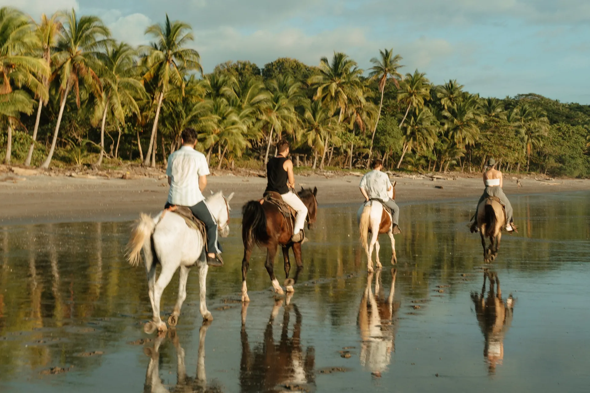 Groep ruiters rijdt te paard langs het strand bij zonsondergang in Santa Teresa, Costa Rica, omringd door palmbomen en weerspiegeling in het water.