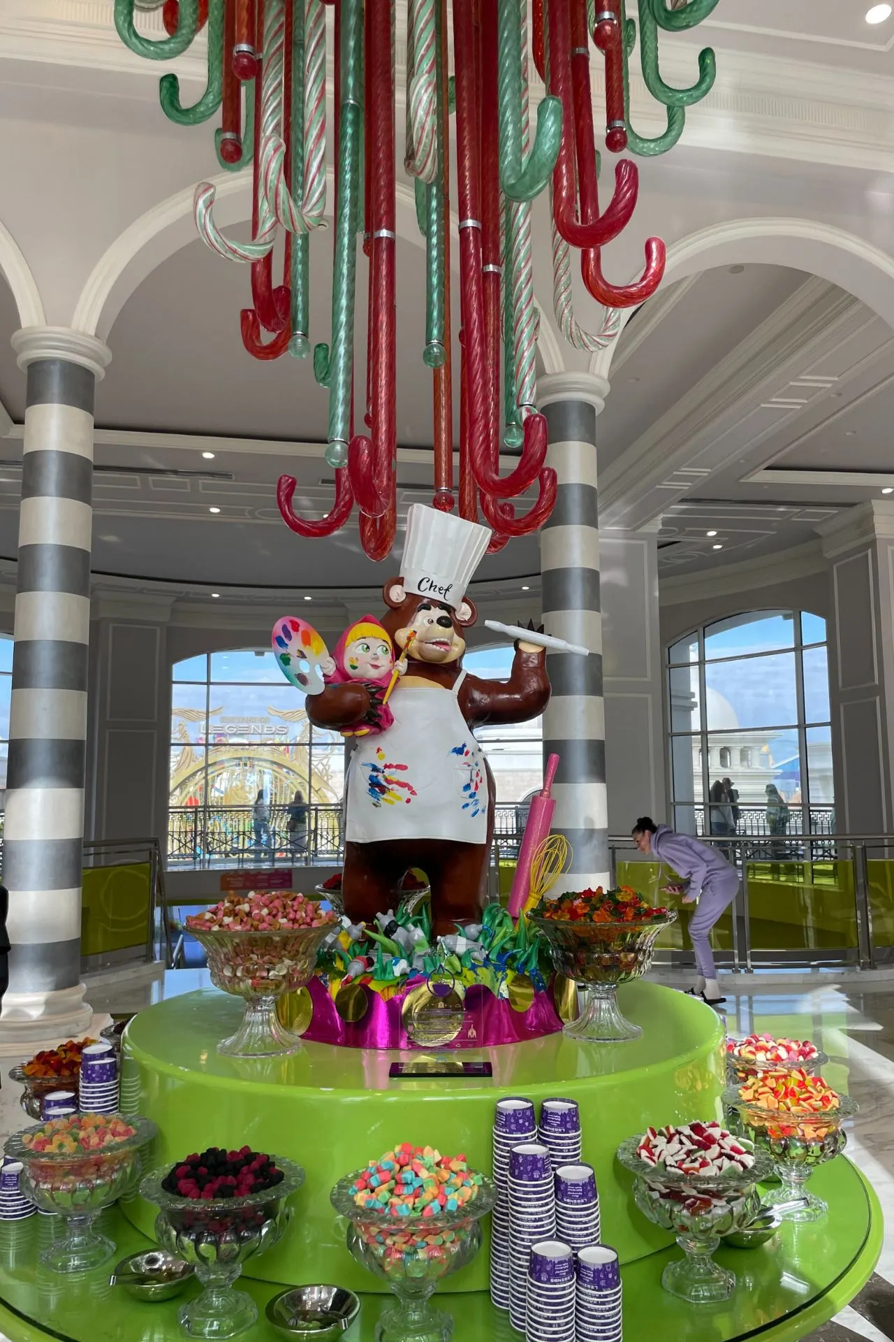 Kleurrijke snoepbar met een groot groen rond display vol glazen potten met snoepjes en een vrolijke berenfiguur als decoratie, omringd door oversized zuurstokken.