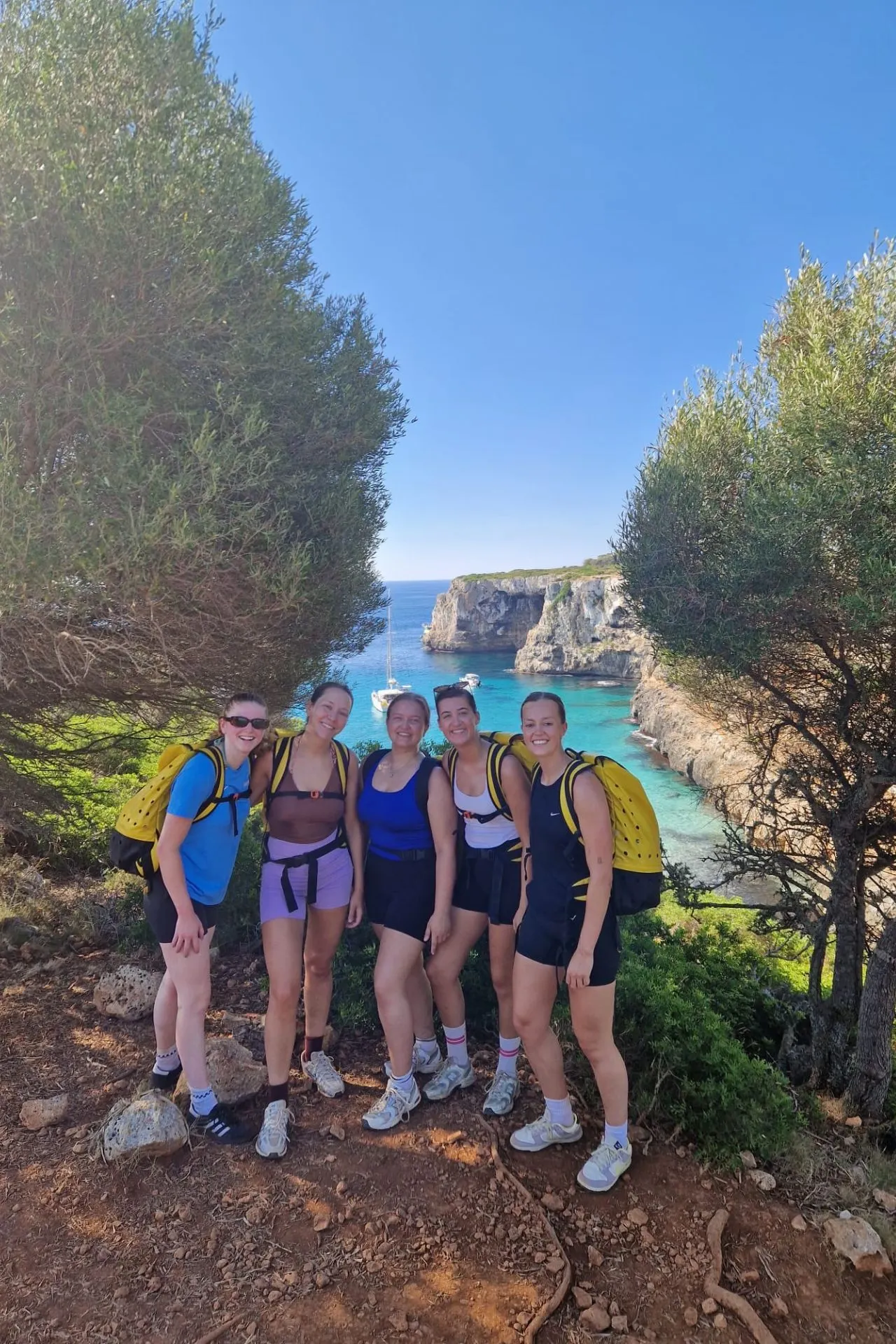 Vijf vrouwen met sportkleding en gele rugzakken poseren lachend op een pad, met op de achtergrond azuurblauw water, kliffen en een zeilboot