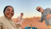 Drie mensen lachen en genieten van een gezellige picknick in de Namibische woestijn. Op de voorgrond staat een tafel met flessen en glazen, terwijl de zandduinen en de heldere lucht de achtergrond vullen.