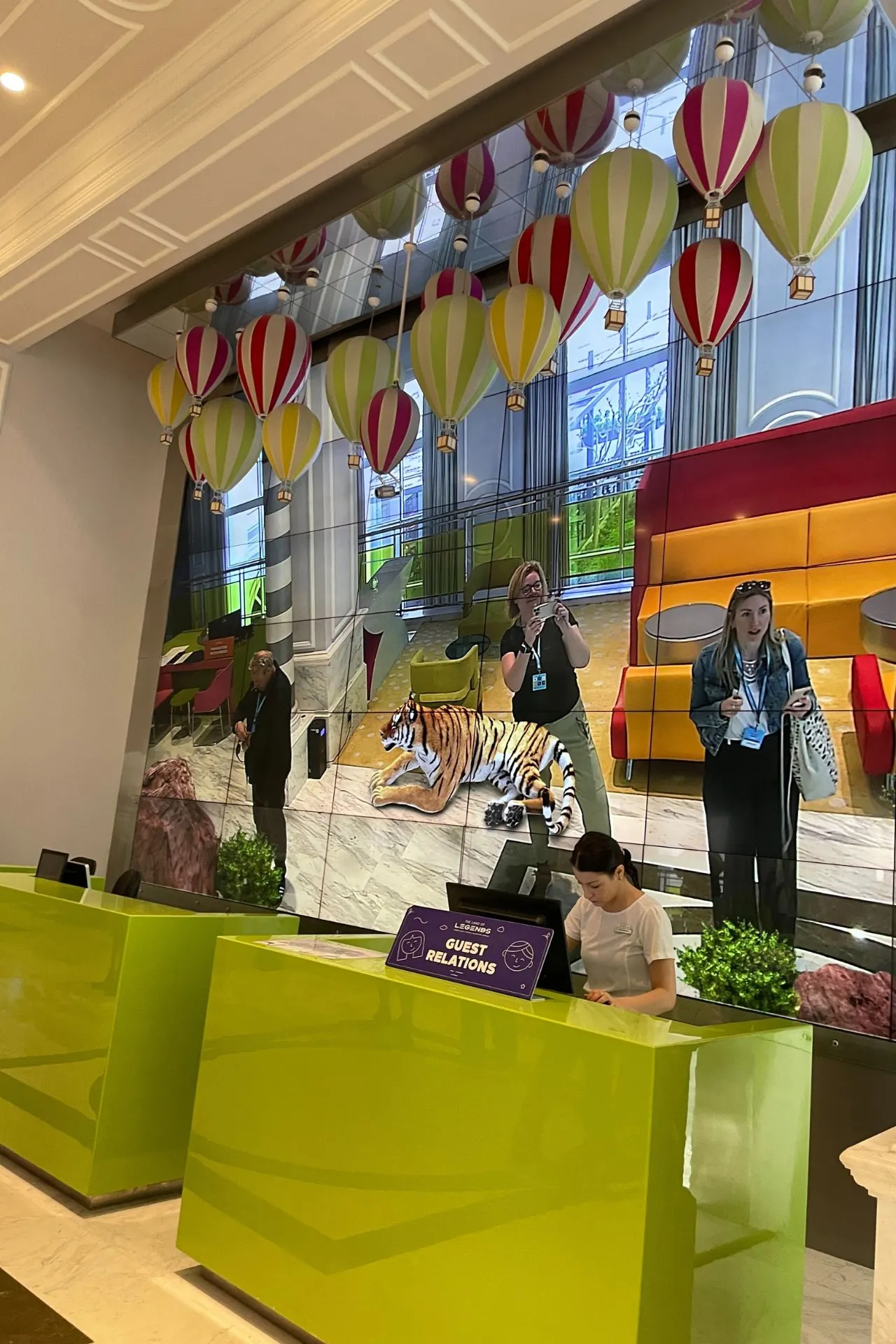 Hotelreceptie met felgroene balies en een grote digitale wand met een kleurrijke animatie van ballonnen en een tijger, waar gasten worden ingecheckt.

