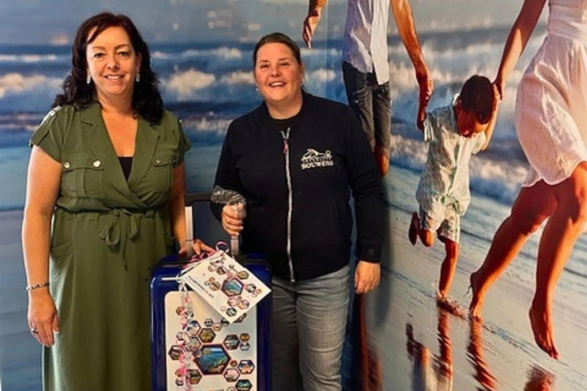 De winnares van de prijsvraag tijdens het KOM Festival in Rosmalen ontvangt een trolley van Travelcenter de Wit. Ze poseert samen met een medewerker van het reisbureau voor een strandfoto op de achtergrond.