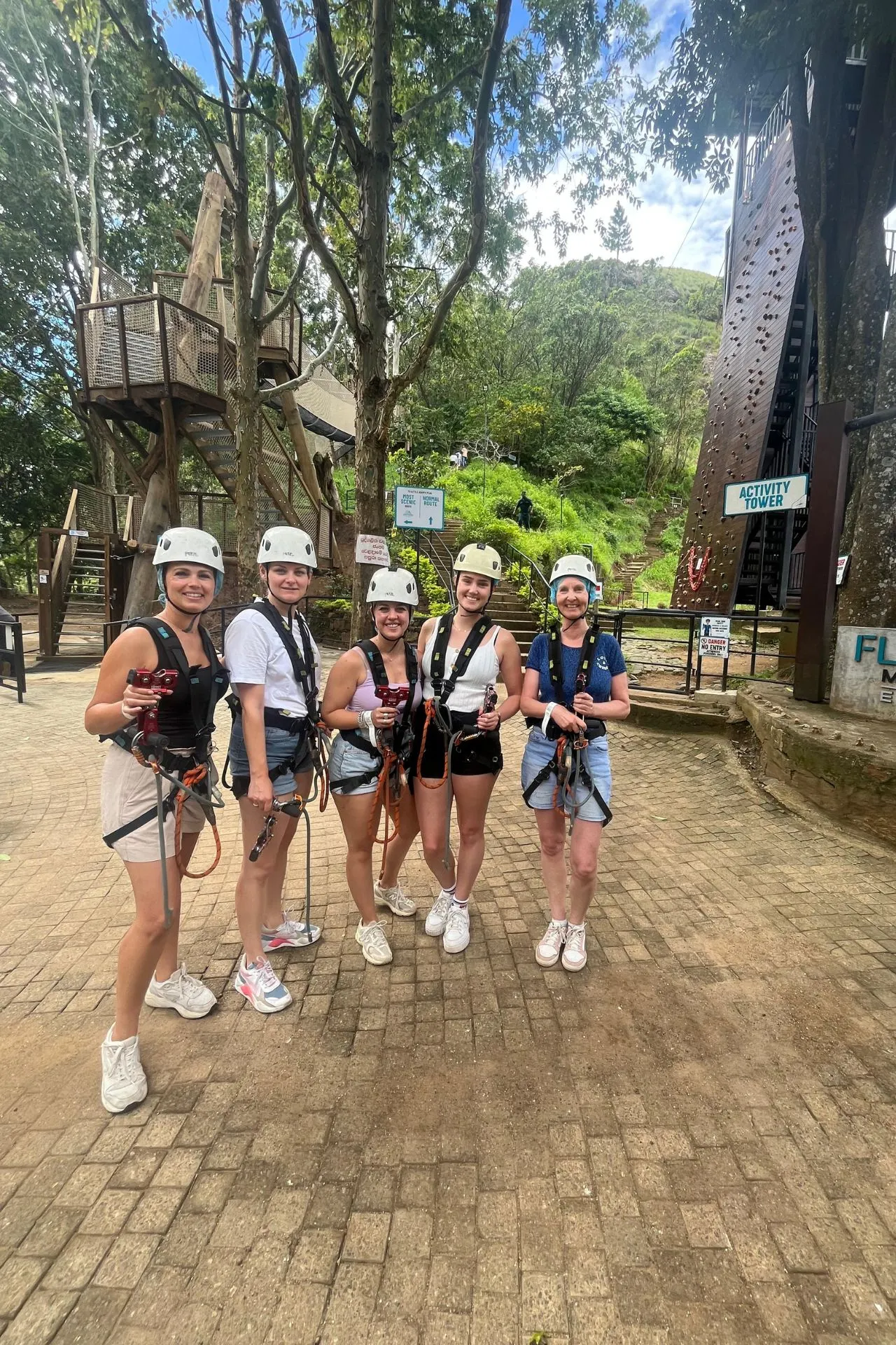 Groepsfoto van vijf vrouwen in sportieve kleding, volledig uitgerust met zipline-harnassen en helmen. Ze staan klaar voor hun rit bij Flying Ravana in Ella, Sri Lanka. Op de achtergrond zijn de toren, trappen en boomrijke omgeving zichtbaar, wat de setting een actieve en natuurlijke sfeer geeft.