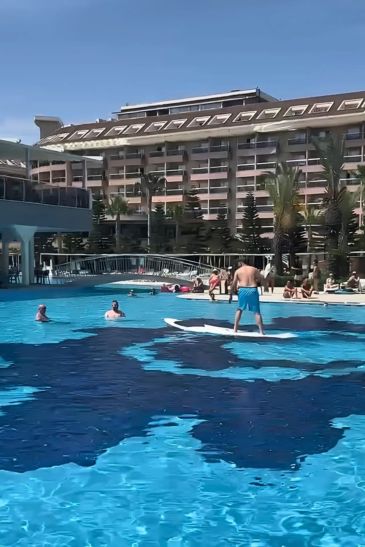 Deelnemers aan de artiestenreis in Turkije doen mee aan een surfchallenge in het grote zwembad van Sunmelia Beach Resort. Op de voorgrond probeert een reiziger in balans te blijven op een surfplank, terwijl andere vakantiegangers toekijken vanaf het water en het zonneterras. Deze sportieve activiteit zorgde voor veel plezier en vormde een leuke afwisseling tussen ontspannen momenten, live muziek en de gezellige feestavonden tijdens de groepsreis aan de Turkse Rivièra.