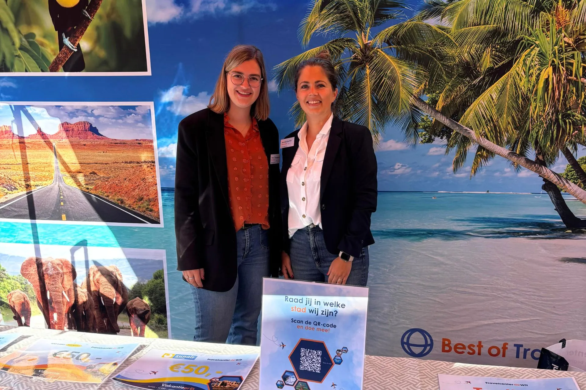 Twee medewerkers van Travelcenter de Wit staan lachend voor een tropische strandachtergrond bij de kraam, naast promotiemateriaal en een bord met een QR-code voor een winactie.