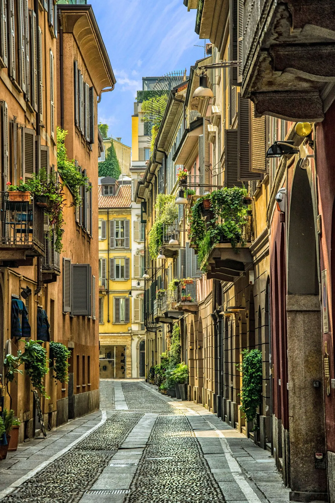 Sfeervolle geplaveide straat in de wijk Brera in Milaan, omringd door kleurrijke gebouwen met houten luiken en balkonnetjes vol groene planten, onder een blauwe hemel.