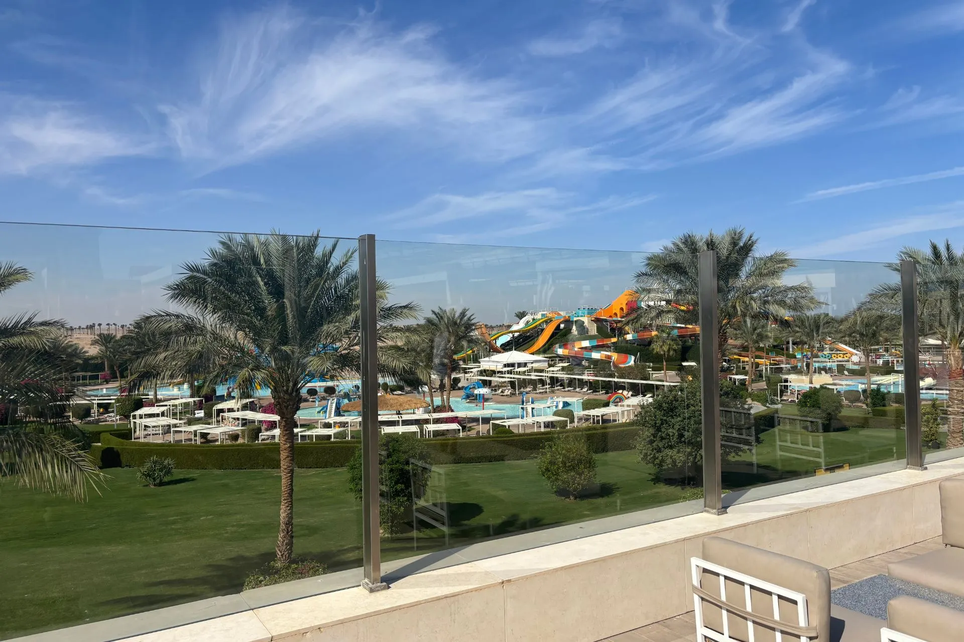 Uitzicht op een waterpark met kleurrijke glijbanen, zwembaden en palmbomen, omgeven door groene grasvelden en een helderblauwe lucht met lichte wolken. Het glazen hek op de voorgrond geeft een moderne uitstraling. Ideaal voor promotie van familievakanties, resorts met aquaparken en actieve ontspanning in Egypte.