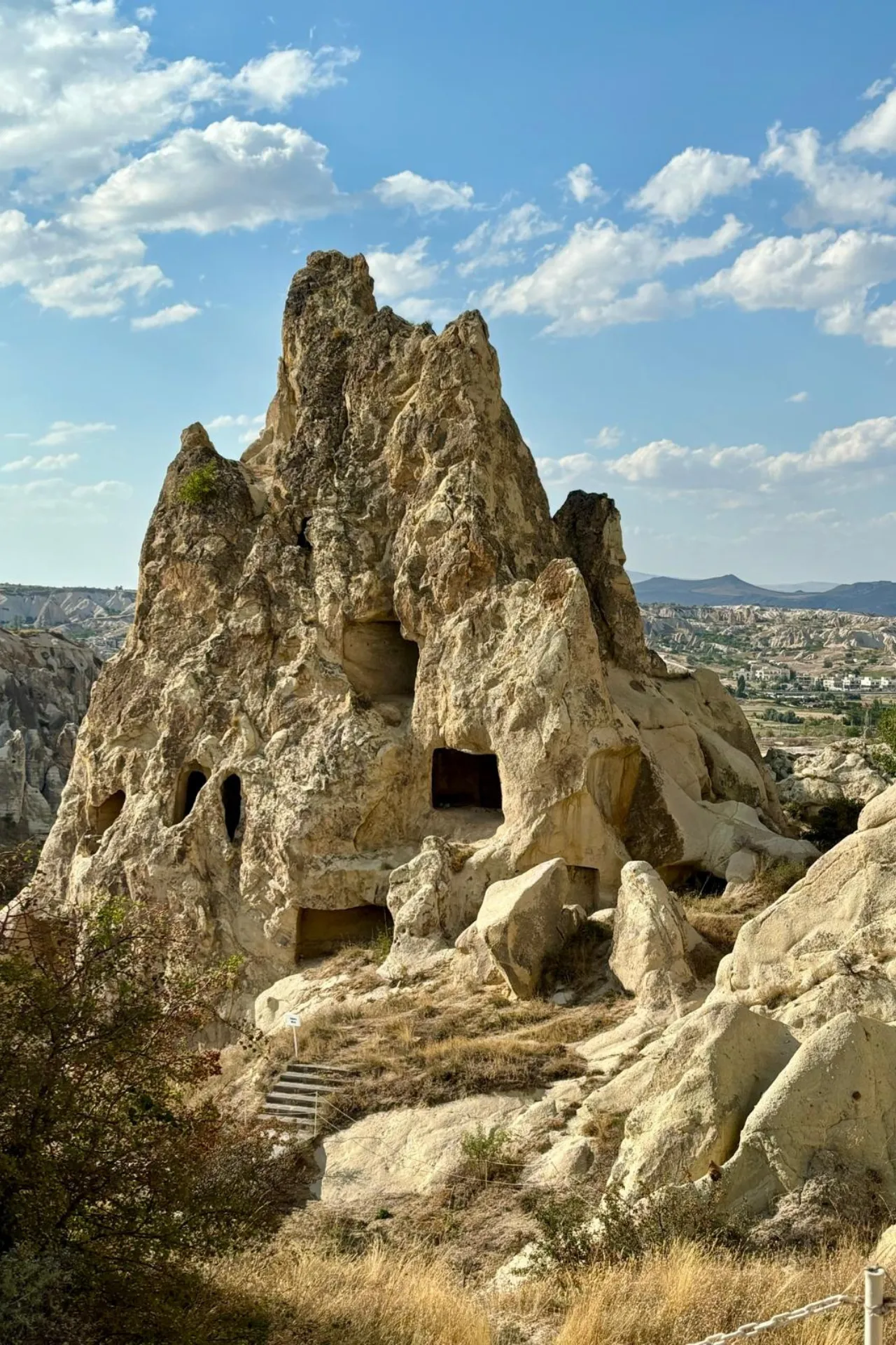 Een indrukwekkende rotsformatie in Cappadocië, Turkije, met uitgehouwen grotten en kamers die eeuwenoude geschiedenis weerspiegelen. De grillige vormen van het zachte vulkanische gesteente contrasteren prachtig met de blauwe lucht en verspreide wolken. Deze iconische rots, omringd door een droog landschap en heuvels in de verte, is een populaire bestemming voor toeristen en fotografen. Perfect voorbeeld van natuurlijke erosie en cultureel erfgoed in het hart van Anatolië, ideaal voor avontuurlijke reizen en unieke bezienswaardigheden.